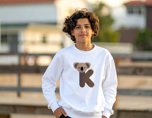 A teenage boy wearing a white crewneck sweatshirt with the letter K in the shape of a Koala illustration, standing in a blurry urban setting.