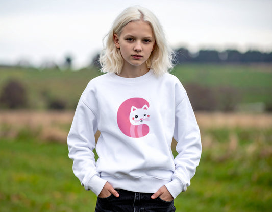 A teenage girl wearing a white crewneck sweatshirt with the letter C in the shape of a cat illustration, standing, blurry countryside background.