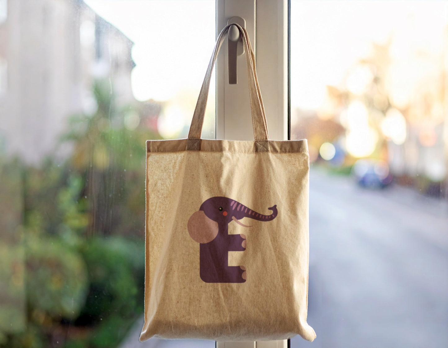 An oyster tote bag with the letter "E" in the shape of an elephant illustration, hanging on a door handle.