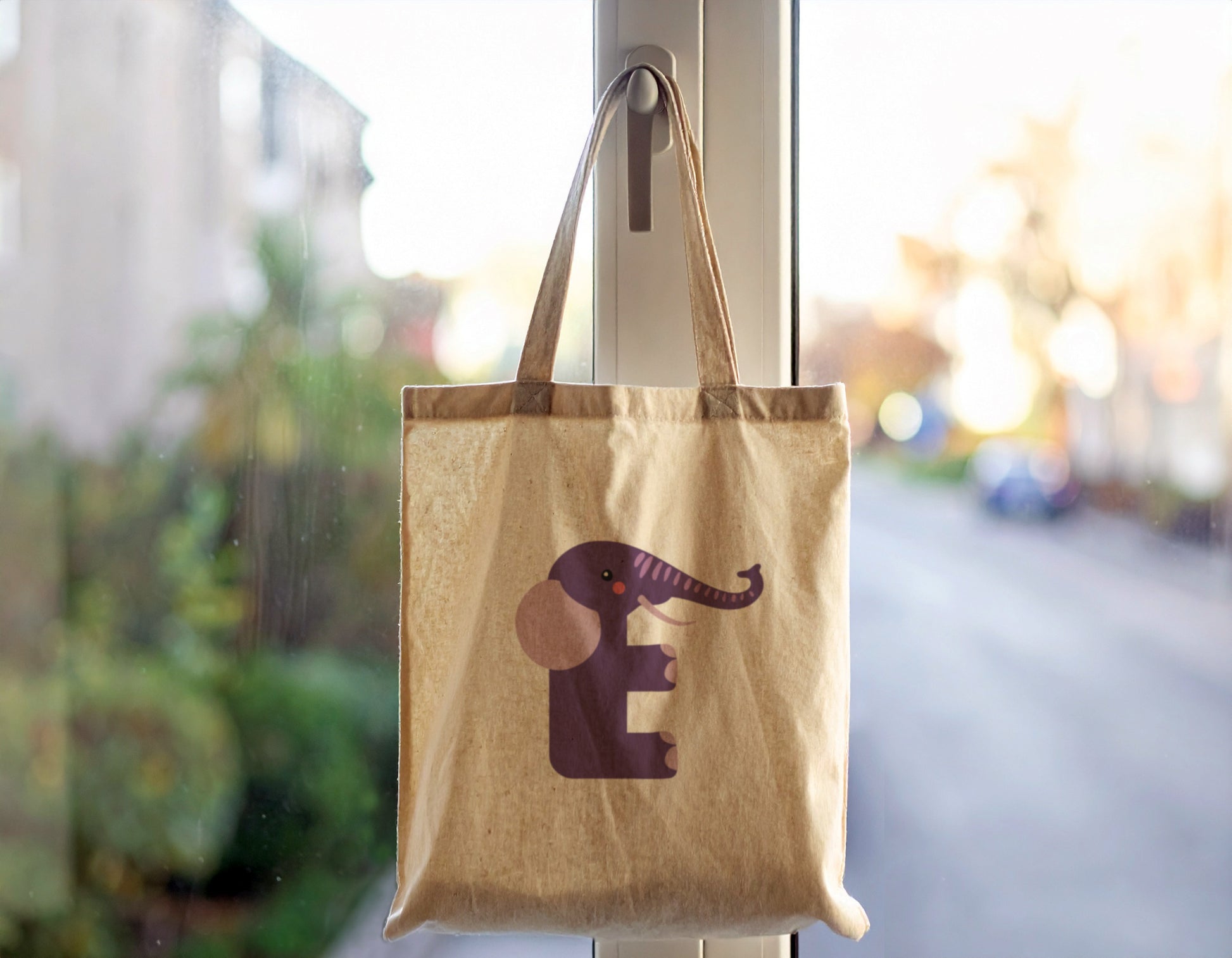 An oyster tote bag with the letter "E" in the shape of an elephant illustration, hanging on a door handle.