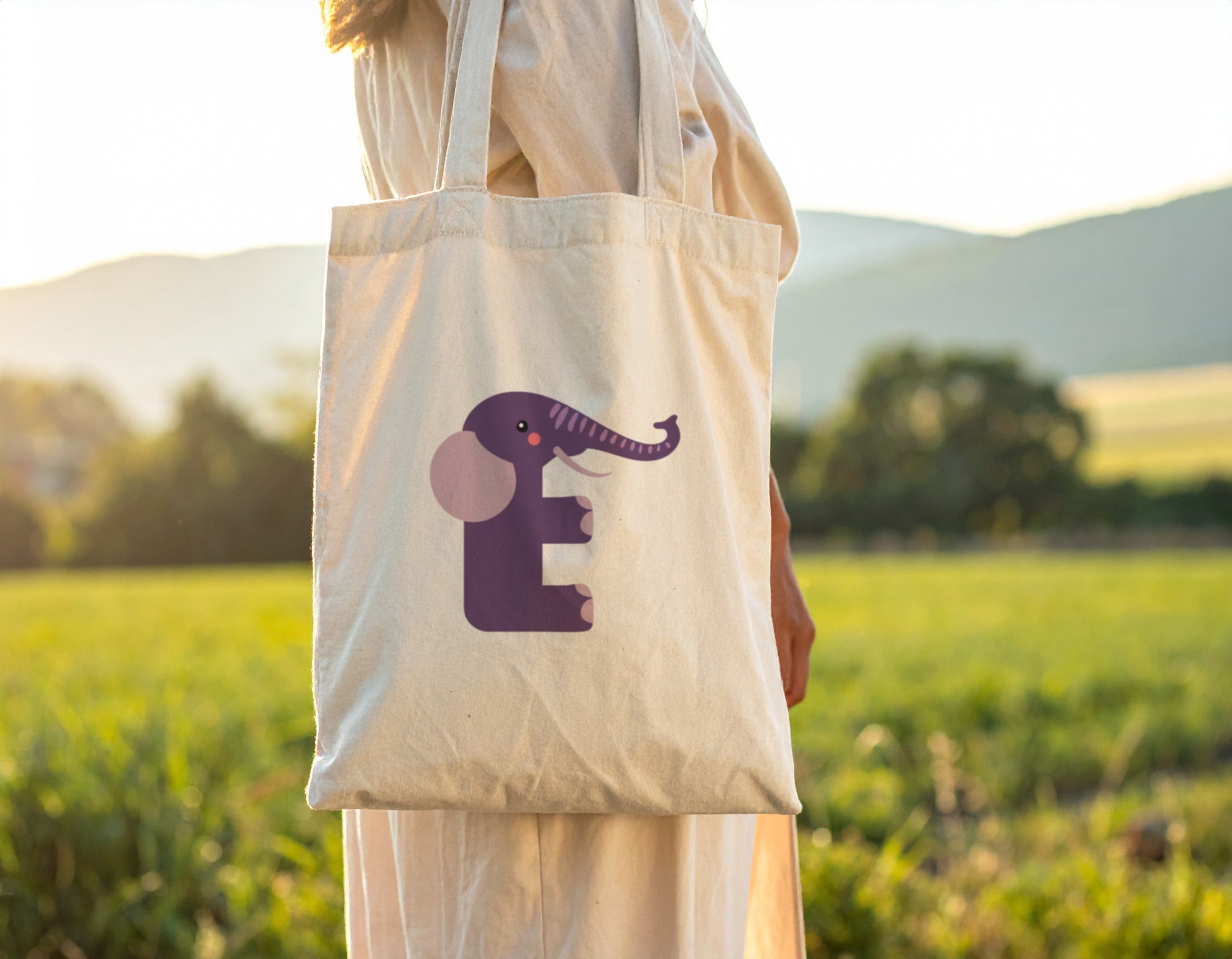 A woman carrying a tote bag with the letter "E" in the shape of an elephant illustration, standing, blurry meadow background.