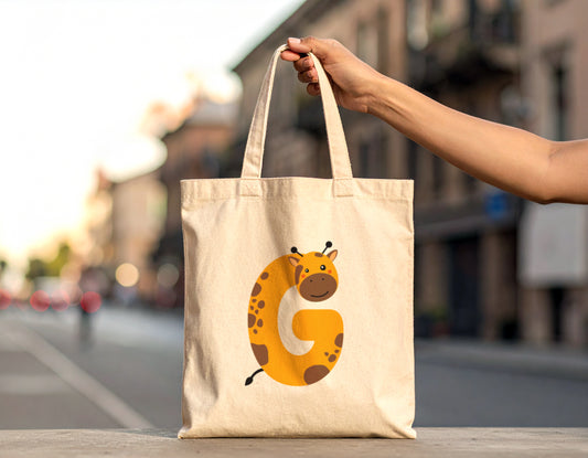 An oyster tote bag with the letter G in the shape of a giraffe illustration held by a woman hand on a blurred city street background.