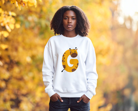 A teenage girl wearing a white crewneck sweatshirt with the letter G in the shape of a giraffe illustration, standing in a blurry autumn background.