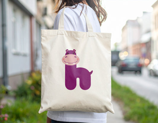 A woman carrying an oyster tote bag with the letter h in the shape of a hippo illustration on a blurred city street background.