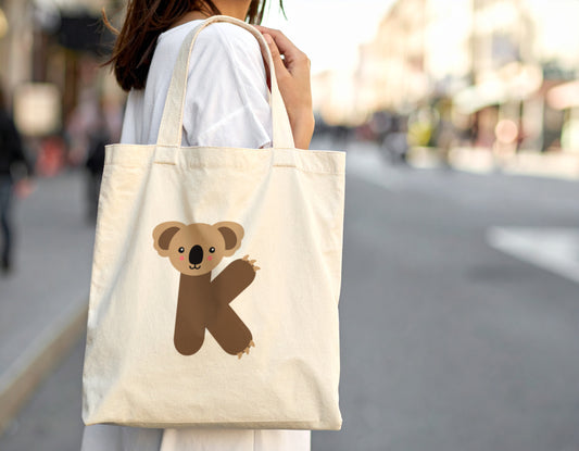 A woman carrying an oyster tote bag with the letter k in the shape of a koala illustration on a blurred city street background.