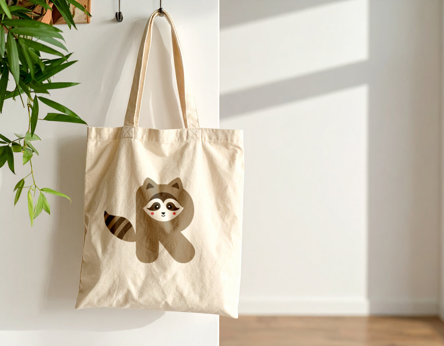 An oyster tote bag with the letter R in the shape of a raccoon illustration, hanging on a hook against a white wall.