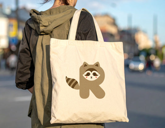 A woman carrying an oyster tote bag with the letter R in the shape of a raccoon illustration on a blurred city street background.