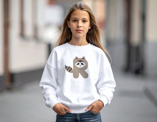 A teenage girl wearing a white crewneck sweatshirt with the letter R in the shape of a raccoon illustration, standing in a blurry school corridor.