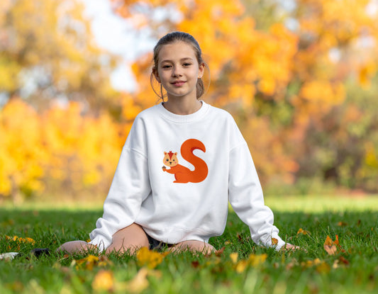 A teenage girl wearing a white crewneck sweatshirt with the letter S in the shape of a squirrel illustration, standing in a blurry autumn background.