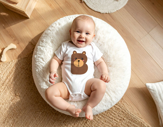 Baby wearing a white organic cotton onesie/bodysuit with the letter B in the shape of a bear illustration, on a white cushion on a wooden floor.