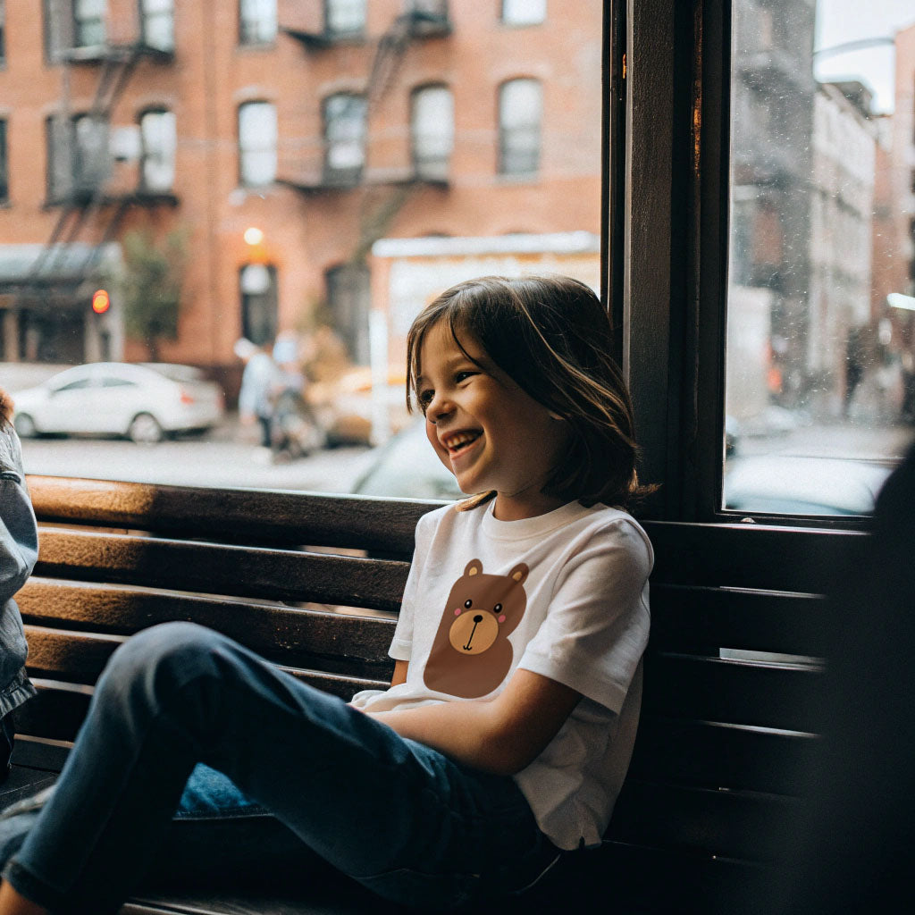 Child wearing a white organic cotton t shirt with the letter B in the shape of a bear illustration, sitting on a bench by a window with a city street view outside.