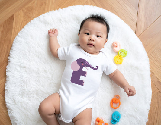 Baby wearing a white organic cotton onesie/bodysuit with the letter E in the shape of an elephant illustration, sitting on a white cushion on a wooden floor.