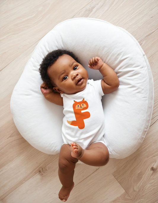 Baby wearing a white organic cotton onesie/bodysuit with the letter F in the shape of a fox illustration, sitting on a white boppy pillow on a wooden floor.
