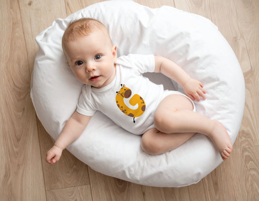 Baby wearing a white organic cotton onesie/bodysuit with the letter G in the shape of a giraffe illustration, sitting on a white boppy pillow on a wooden floor.