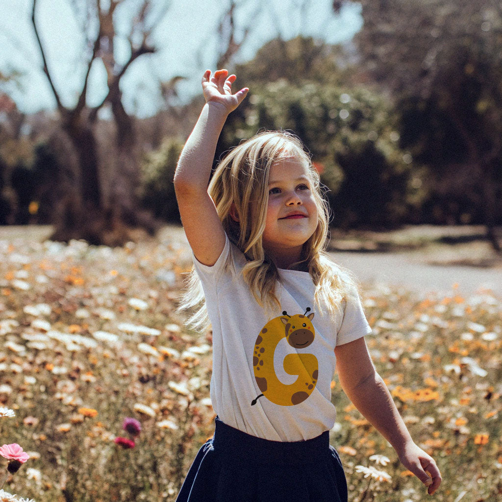 Cute girl child in a field of flowers wearing a t shirt with the letter G in the shape of a giraffe illustration.