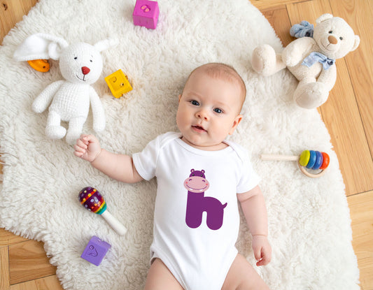 Baby wearing a white organic cotton onesie/bodysuit with the letter H in the shape of a purple hippo illustration, lying on a fluffy white carpet on a wooden floor.