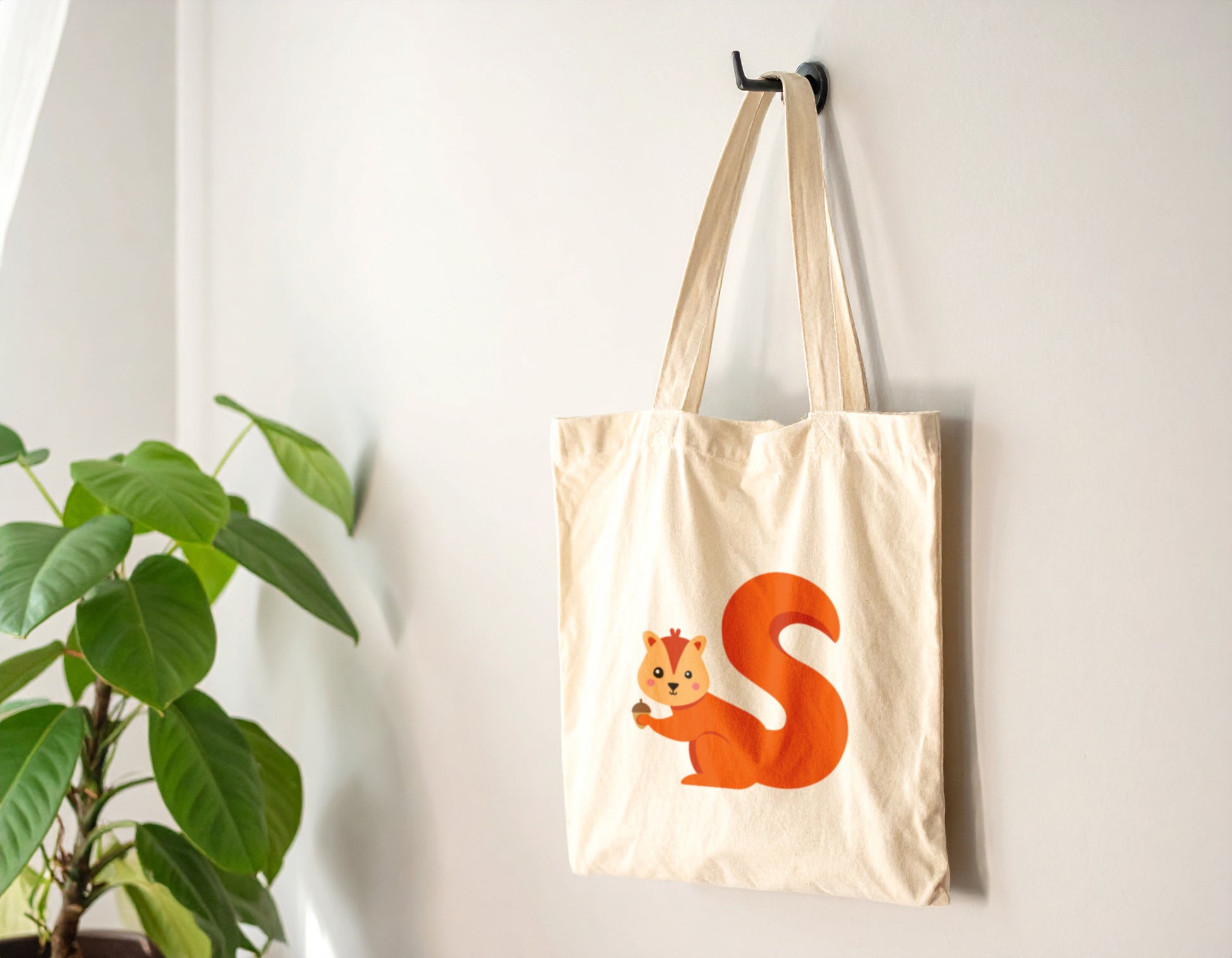 An oyster tote bag with the letter "S" in the shape of a squirrel illustration, hanging on a wooden hook against a white wall.